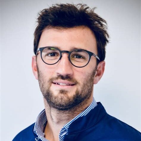 Head-and-shoulders portrait of a man with dark hair and round glasses, wearing a blue jacket and striped shirt, looking at the camera.