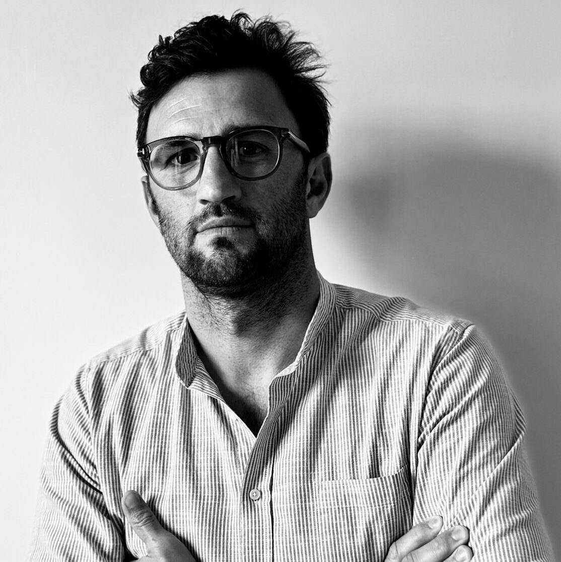 Man with dark hair and beard wearing glasses and a striped shirt, arms crossed, looking at the camera.
